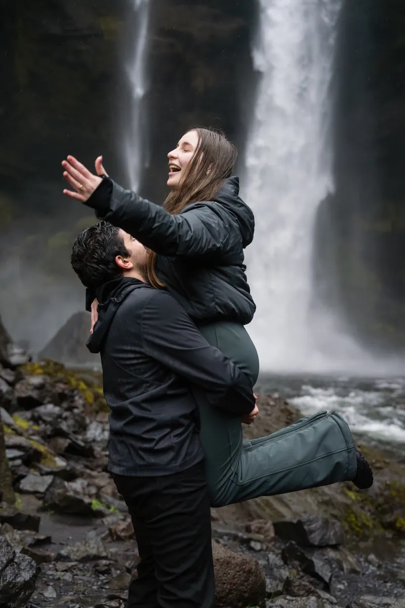 Edu levanta en brazos a Dani, que echa la cabeza atrás y abre los brazos riendo, frente a la cascada.